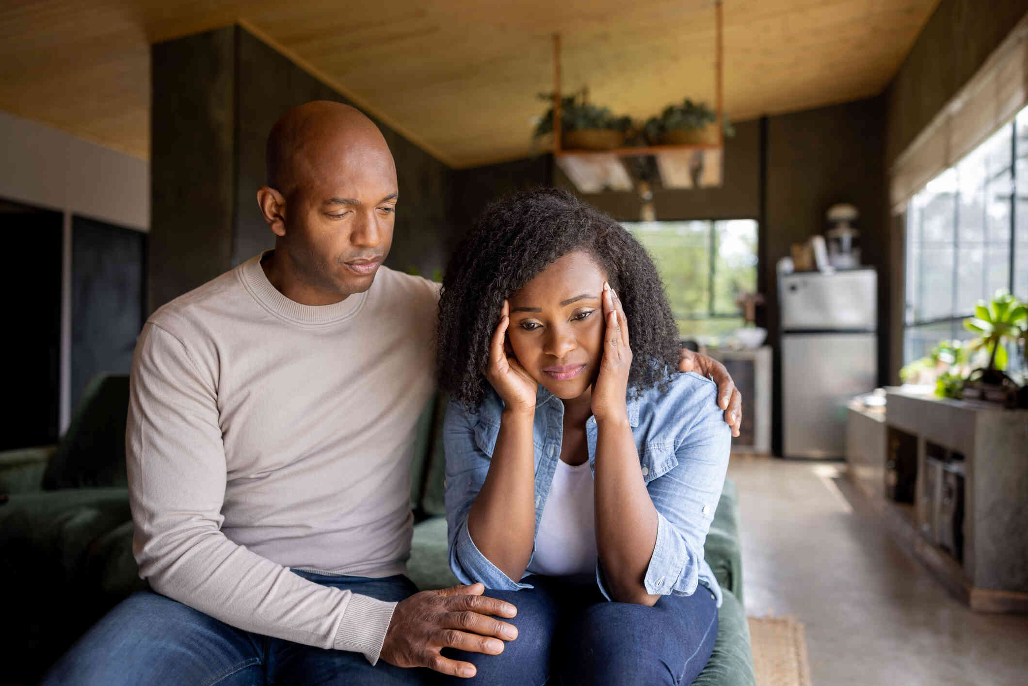 A bald man wraps his arm around a woman sitting next to him. The woman has curly hair and a pained expression as she massages the side of her face with her hands.